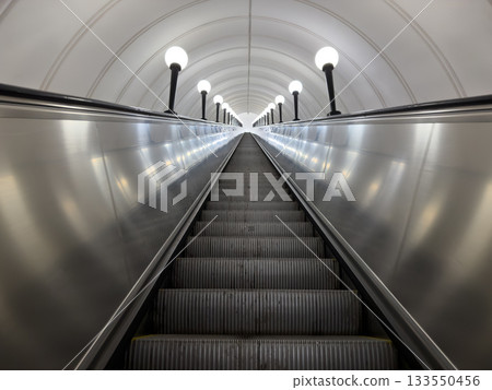 Massive metal escalator leading upward from subway underpass. Numerous steps empty of people. Riding escalator up along path illuminated by lanterns. Tunnel roof round with escalator Massive metal escalator leading upward from subway underpass. Numerous steps empty of people. Riding escalator up along path illuminated by lanterns. Tunnel roof round with escalator 133550456