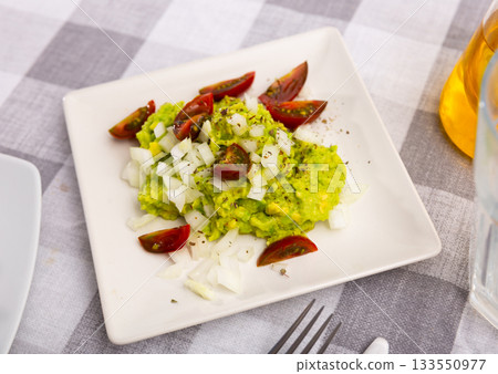 Pile of guacamole on plate, sprinkled with chopped hot onions, decorated with slices of tomatoes 133550977