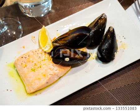 Piece of fried salmon is served on rectangular plate, and decorated with mussels. Piece of fried salmon is served on rectangular plate, and decorated with mussels. 133551083