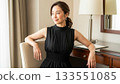 A smiling Japanese woman in a black dress sitting in a desk chair in a hotel 133551085