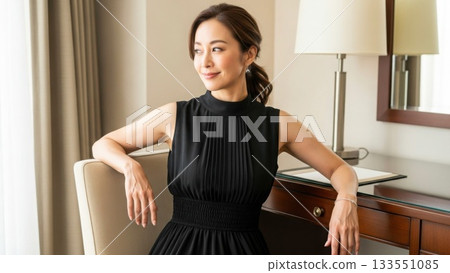A smiling Japanese woman in a black dress sitting in a desk chair in a hotel 133551085