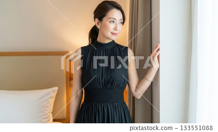 Elegant woman in a black dress standing by a hotel window looking out the window Elegant woman in a black dress standing by a hotel window looking out the window 133551088