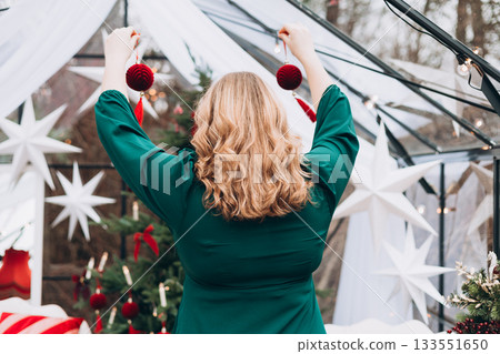 Young woman in green dress decorating Christmas tree at home and smiling. Winter time and holidays. Celebration banner. back view Young woman in green dress decorating Christmas tree at home and smiling. Winter time and holidays. Celebration banner. back view 133551650