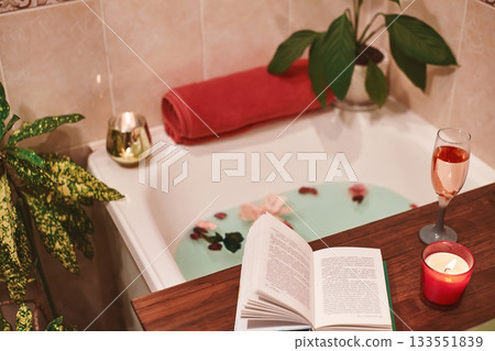 Bath with flower petals. Book, candles and glass of wine on a wood tray. Organic Spa Relaxation in comfort cozy bathroom Bath with flower petals. Book, candles and glass of wine on a wood tray. Organic Spa Relaxation in comfort cozy bathroom 133551839
