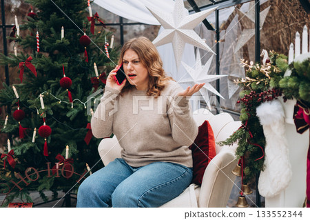 Young angry blonde woman talking on smartphone celebrating Christmas near decorated Christmas tree. 133552344