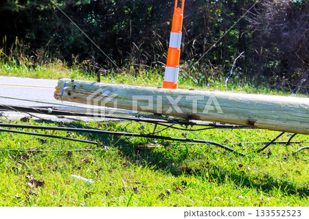 Broken utility pole lies on grass by road, marked by an orange cone in recent hurricane storm 133552523
