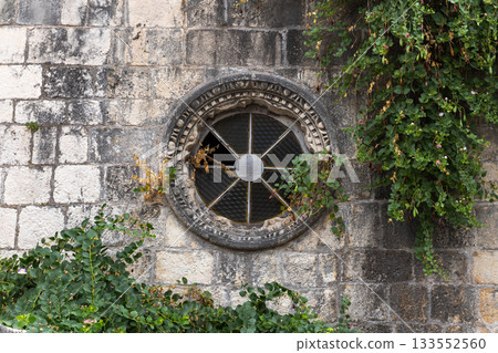 An ancient stone wall features a round, iron-grated window framed by carved stone 133552560