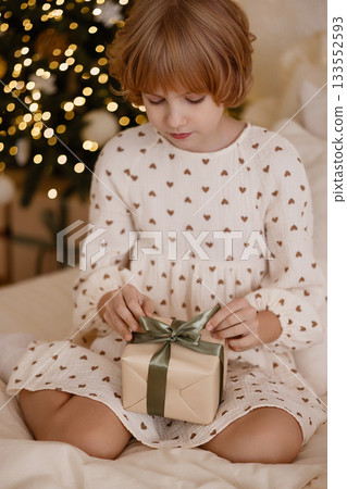 Young girl holding gift on bed in cozy holiday room with Christmas tree and white wall 133552593