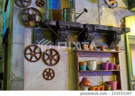 Wooden shelves display terracotta pots, colorful buckets, and a metal watering can beside stained glass. Night lighting reveals textures, gears, and handcrafted decor. Wooden shelves display terracotta pots, colorful buckets, and a metal watering can beside stained glass. Night lighting reveals textures, gears, and handcrafted decor. 133553167