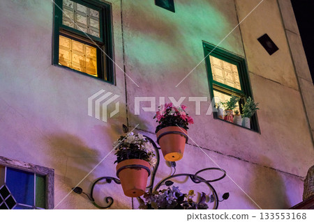 Terracotta planters with flowers hang on a lighted stucco facade with small windows. Soft night colors highlight the decorative urban scene. Terracotta planters with flowers hang on a lighted stucco facade with small windows. Soft night colors highlight the decorative urban scene. 133553168