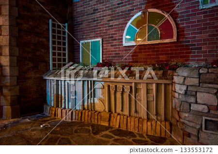 Flower bed against a red brick wall beneath a colorful arched window. Night lighting reveals blossoms, wood textures, and decorative details. 133553172