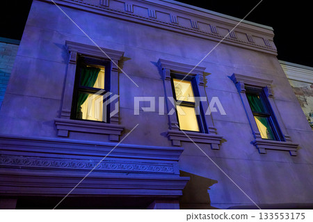 Neoclassical facade with three framed windows and green curtains glowing from inside. Evening illumination reveals stone moldings and clean symmetry. 133553175