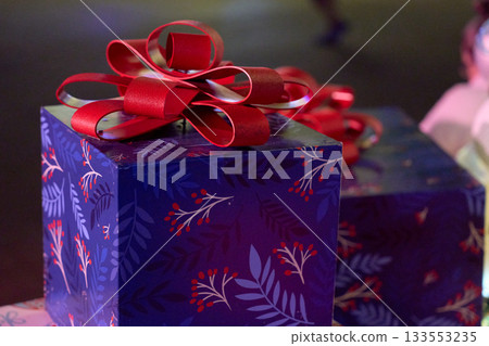 A blue gift box with a large red ribbon bow sits under holiday lights. The wrapped present shows festive colors and glossy paper in a closeup night scene. A blue gift box with a large red ribbon bow sits under holiday lights. The wrapped present shows festive colors and glossy paper in a closeup night scene. 133553235