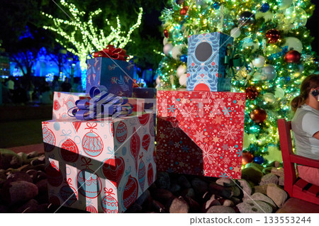 Stacks of wrapped presents with bows sit by a glowing Christmas tree. Holiday lights and ornaments create a bright, festive night scene. 133553244