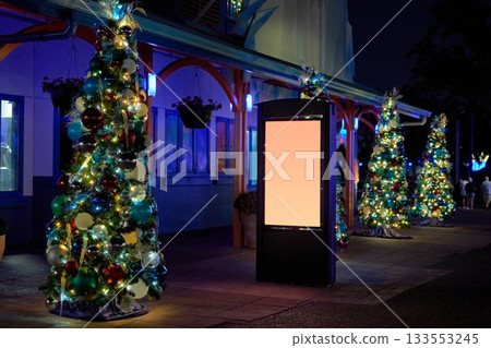 Multiple small Christmas trees with shiny ornaments line the path near a storefront. A blank digital sign stands among the glowing holiday lights. 133553245