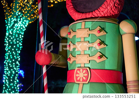 A closeup view shows the green jacket, red belt, and candy cane staff of a giant Elmo nutcracker statue. Holiday lights on nearby trees add colorful bokeh in the night background. A closeup view shows the green jacket, red belt, and candy cane staff of a giant Elmo nutcracker statue. Holiday lights on nearby trees add colorful bokeh in the night background. 133553251