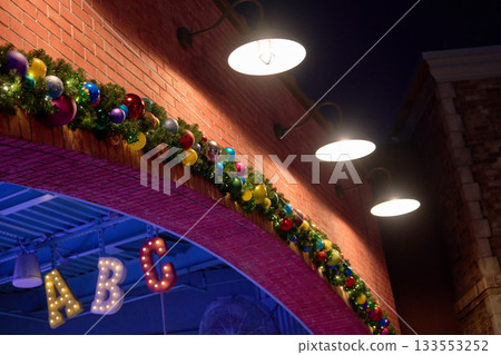 A brick archway is decorated with colorful Christmas garland and shiny ornaments under glowing lamps. Lit letters A, B, and C hang inside the entrance in a festive nighttime scene. 133553252