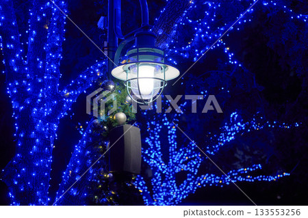 Lamp post wrapped in garland and ornaments glows at night. Blue Christmas lights sparkle on trees creating a festive scene. 133553256