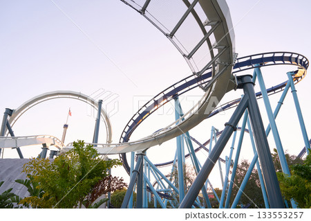 Curving coaster tracks weave through blue columns at dusk. Soft sky and plants frame a dynamic theme park scene. Curving coaster tracks weave through blue columns at dusk. Soft sky and plants frame a dynamic theme park scene. 133553257