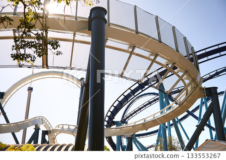 Curving coaster rails pass under a safety net with palms and blue sky. Sunlight and steel shapes create a lively theme park view. 133553267