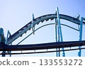 A steel roller coaster climbs a tall curve with riders suspended beneath the track. The amusement ride stands out against a bright blue sky. 133553272