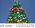 The top of a Christmas tree is covered with many bright baubles in red, gold, and blue. The festive holiday decoration stands tall against a clear blue sky. 133553278