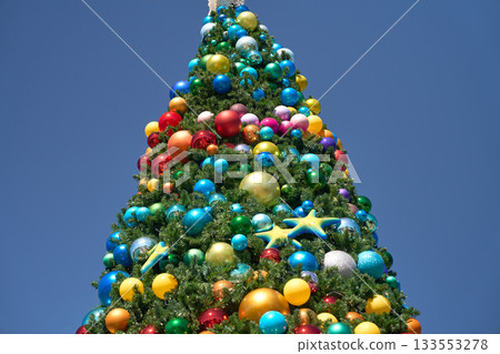 The top of a Christmas tree is covered with many bright baubles in red, gold, and blue. The festive holiday decoration stands tall against a clear blue sky. 133553278