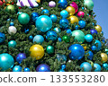 Closeup view of a Christmas tree covered with large colorful baubles in blue, green, and gold. Shiny ornaments sparkle in the sun on dense evergreen branches outdoors. 133553280