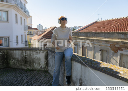 Woman sitting on urban rooftop listening music with headphones Woman sitting on urban rooftop listening music with headphones 133553351