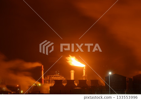 An industrial flare stack burns off excess gas with a large, bright orange flame and thick smoke 133553996