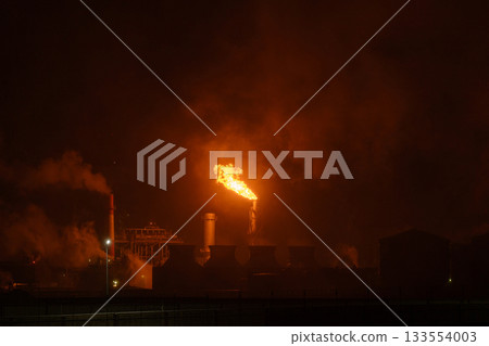 An industrial flare stack burns off excess gas with a large, bright orange flame and thick smoke 133554003