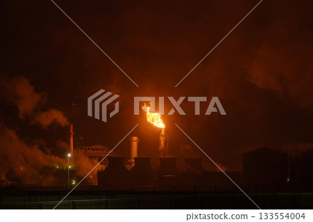 An industrial flare stack burns off excess gas with a large, bright orange flame and thick smoke 133554004