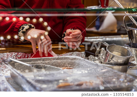 Market vendor in a red sweater preparing fried pastry behind a glass counter at a winter holiday market. Foil trays, metal tools and warm festive lights create a cozy Christmas atmosphere Market vendor in a red sweater preparing fried pastry behind a glass counter at a winter holiday market. Foil trays, metal tools and warm festive lights create a cozy Christmas atmosphere 133554853