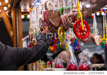 Customer hand selecting an item at a vibrant display of colorful candy wheels, gingerbread hearts, and festive sweets at a traditional christmas market stall Customer hand selecting an item at a vibrant display of colorful candy wheels, gingerbread hearts, and festive sweets at a traditional christmas market stall 133554855