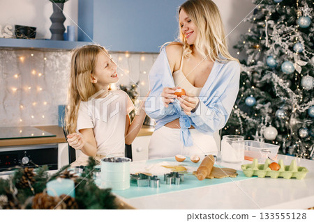 Family preparation of ginger biscuits with daughter. Mother and little girl break an egg. Blonde woman wearing blue shirt and girl white t-shirt. 133555128