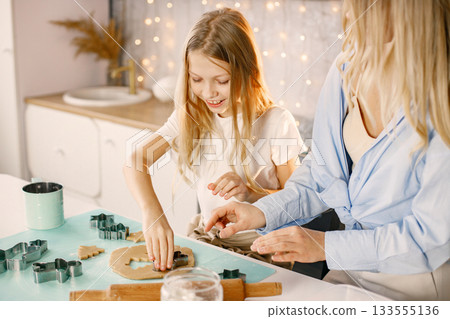 Family preparation of ginger biscuits with daughter. Mother and little girl cooking cookies. Blonde woman wearing blue shirt and girl white t-shirt. Family preparation of ginger biscuits with daughter. Mother and little girl cooking cookies. Blonde woman wearing blue shirt and girl white t-shirt. 133555136