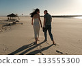 Couple walking barefoot on sandy beach near ocean at sunset, wind blowing her hair in a peaceful romantic scene. 133555300