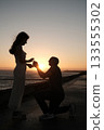 Man proposing to a woman at sunset near the ocean, romantic silhouette moment captured on a seaside promenade. 133555302