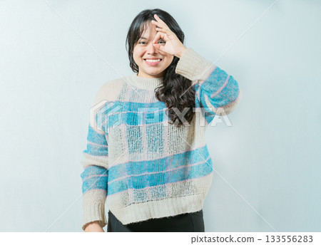 Beautiful asian woman making OK gesture looking through fingers isolated. Smiling asian girl making gesturing OK looking through fingers 133556283