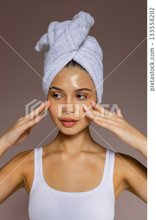 Young woman massaging her face with both hands after a shower, wearing a towel on her head and showing a fresh skincare routine. Young woman massaging her face with both hands after a shower, wearing a towel on her head and showing a fresh skincare routine. 133558202