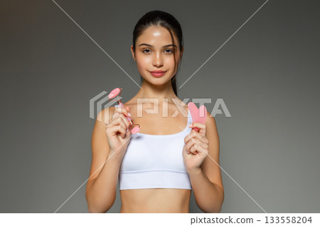 Young woman holding a pink facial roller and a gua sha stone, presenting skincare massage tools during a beauty routine on a neutral studio background. Young woman holding a pink facial roller and a gua sha stone, presenting skincare massage tools during a beauty routine on a neutral studio background. 133558204