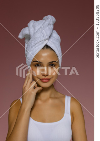 Young woman applying skincare cream to her cheek after a shower, wearing a towel on her head and showing a gentle beauty routine. 133558209