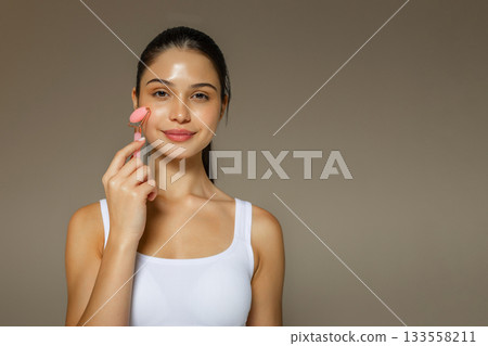 Young woman using a pink facial roller on her cheek, demonstrating a relaxing skincare massage routine on a soft studio background. Young woman using a pink facial roller on her cheek, demonstrating a relaxing skincare massage routine on a soft studio background. 133558211