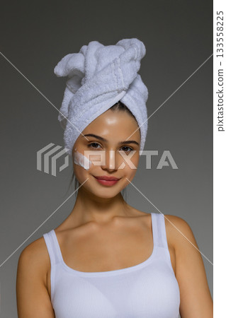 Young woman applying skincare cream to her cheek after a shower, wearing a towel on her head and showing a gentle beauty routine. Young woman applying skincare cream to her cheek after a shower, wearing a towel on her head and showing a gentle beauty routine. 133558225