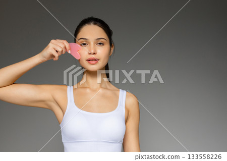 Young woman gently massaging her face with a pink gua sha stone, demonstrating a skincare routine against a neutral background. 133558226