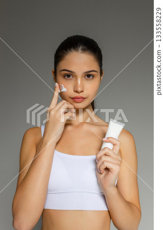 Young woman applying face cream while holding a cosmetic tube, demonstrating a hydrating skincare step on a clean studio background. 133558229