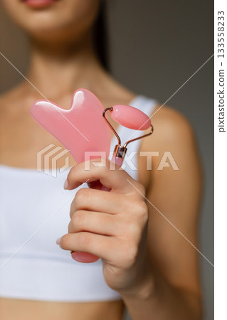 Woman holding a pink facial roller and a gua sha stone, demonstrating skincare massage tools on a neutral background. Woman holding a pink facial roller and a gua sha stone, demonstrating skincare massage tools on a neutral background. 133558233