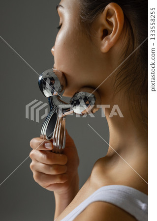 Close-up of a young woman massaging her neck with a metal 3D roller, demonstrating a tightening and relaxing skincare routine at home. 133558255