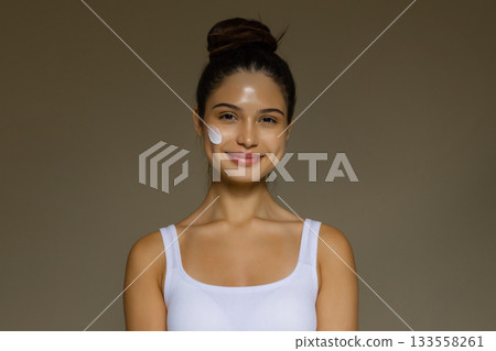 Young woman smiling with a skincare cream applied to her cheek, demonstrating a daily beauty routine. Young woman smiling with a skincare cream applied to her cheek, demonstrating a daily beauty routine. 133558261