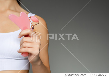Woman holding a pink gua sha stone and a facial roller, demonstrating skincare tools on a neutral background. Woman holding a pink gua sha stone and a facial roller, demonstrating skincare tools on a neutral background. 133558277
